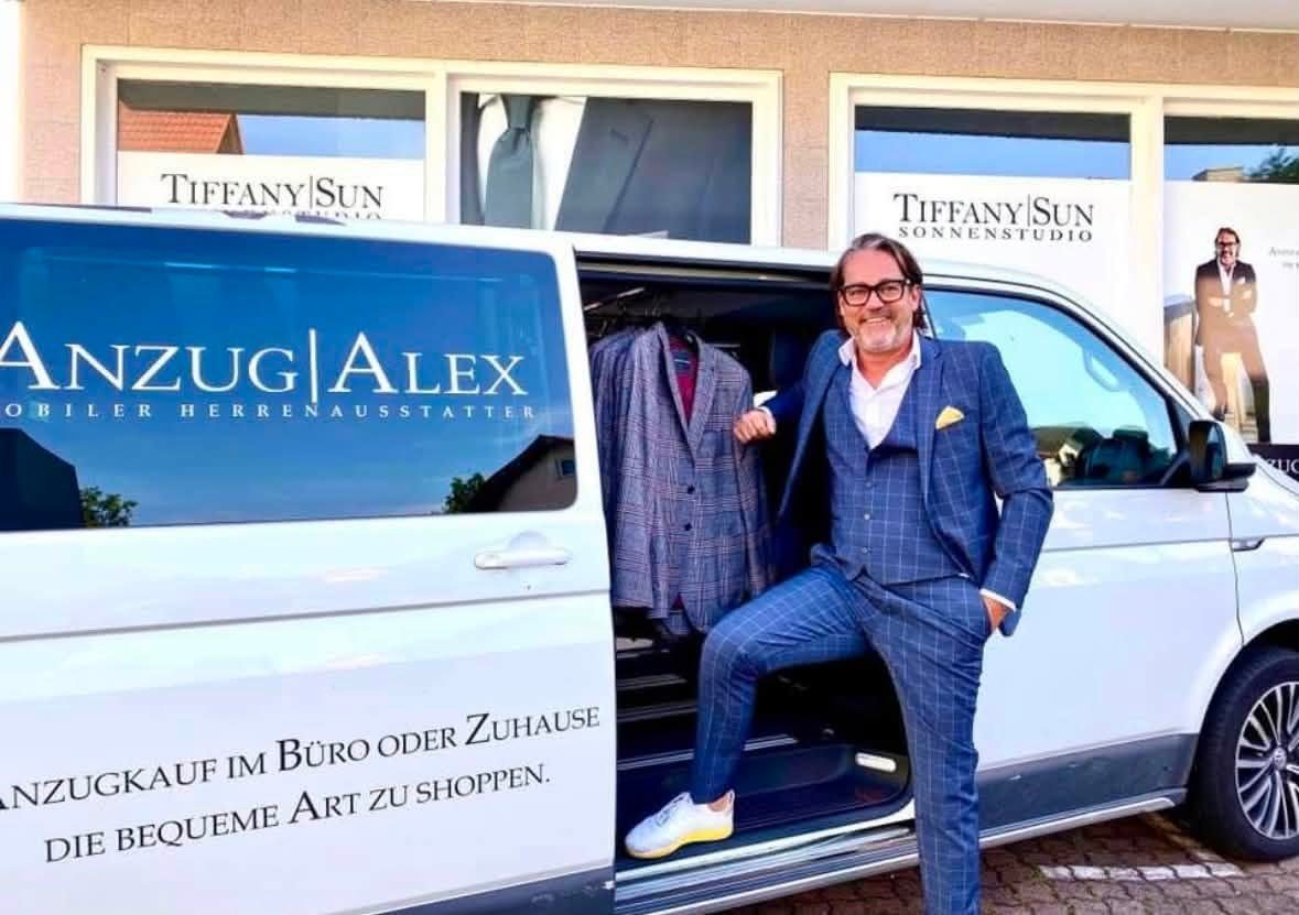 Alexander Desch im blau karierten Anzug an der offenen Schiebetür seines weißen Anzug-Alex-Busses voller Sakkos