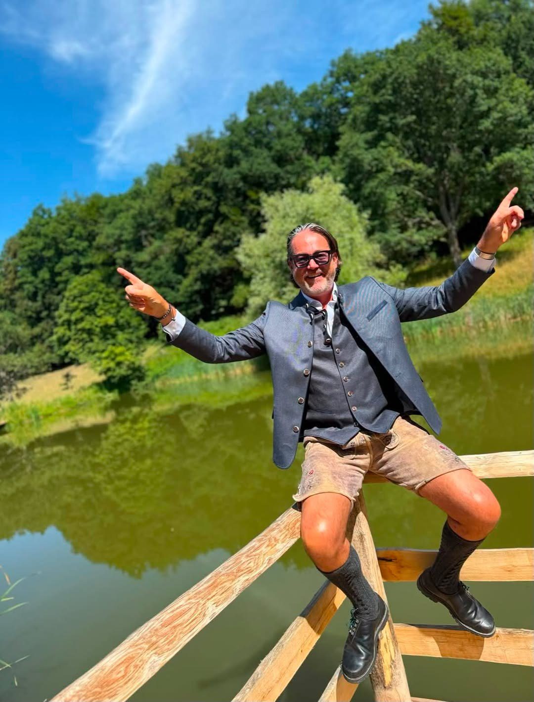 Alexander Desch gut gelaunt in Trachtenjanker und Lederhose auf einem Holzgeländer am See